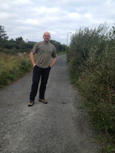 The Independent county councillor stands at some overgrown hedges.